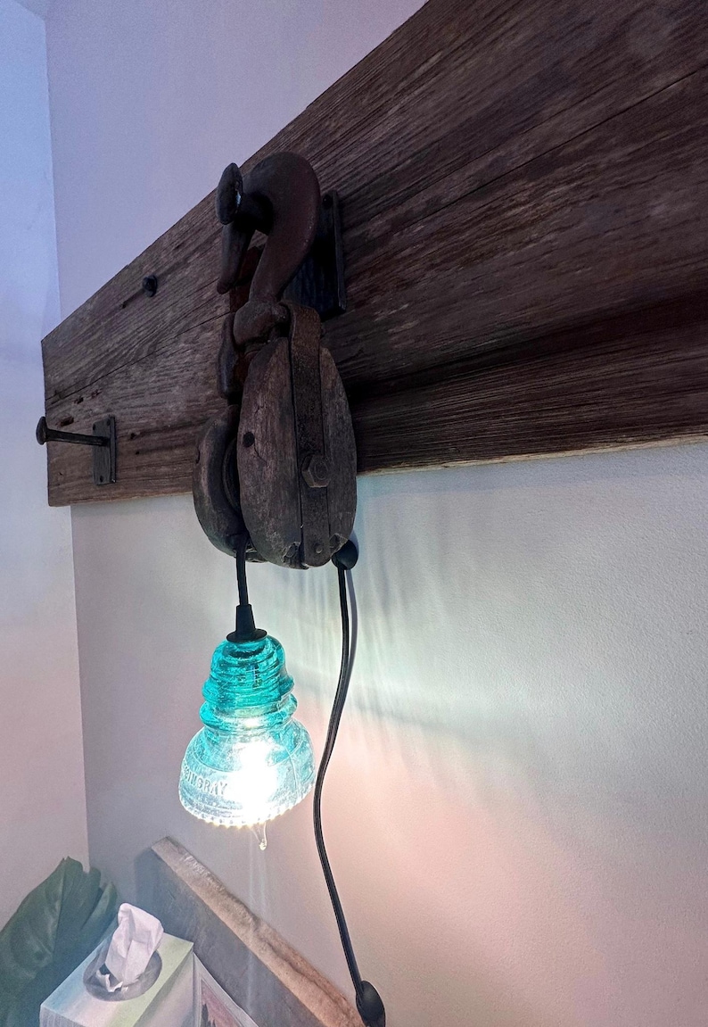 May include: A wall-mounted light fitting featuring a weathered wooden board with a dark metal hook and a vintage wooden pulley block. A bright, translucent turquoise glass insulator serves as the lampshade, suspended by a black electrical cord.