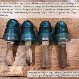 May include: Four vintage glass insulators in varying shades of teal and blue, each mounted on a weathered, cylindrical wooden post. The insulators feature a rounded top and a flared base, displaying a smooth, translucent surface texture.