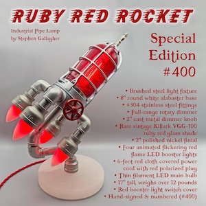 May include: A unique industrial pipe lamp in the shape of a rocket, featuring a red glass shade and animated flickering red flame LED booster lights. The lamp is 17 inches tall and has a white alabaster base. The text "RUBY RED ROCKET" is at the top.