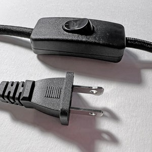 May include: A black electrical power cord featuring a rectangular inline rocker switch with a single black button and a standard two-prong polarized plug. The cord is covered in a black fabric weave, and the switch housing is made of black plastic.