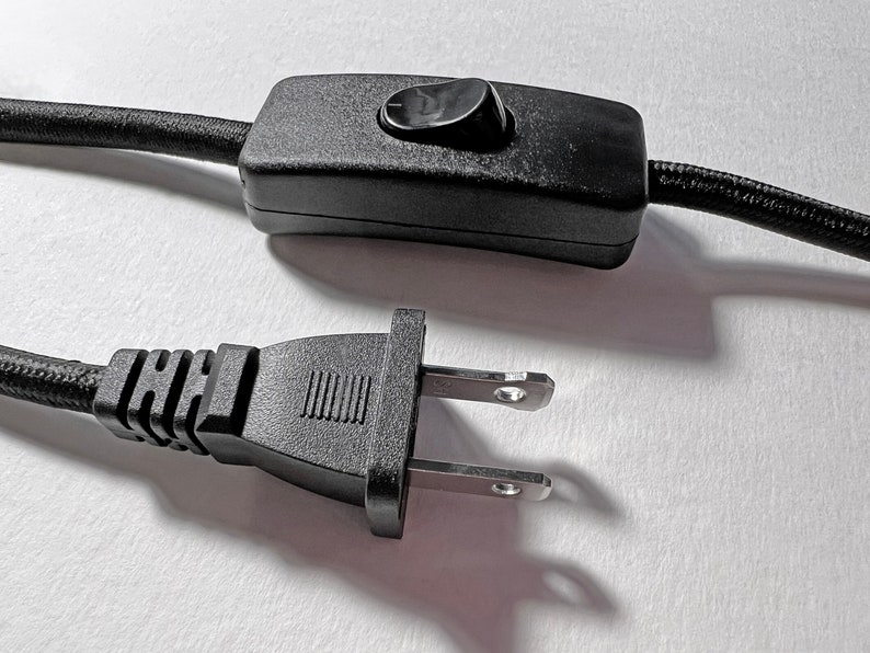 May include: A black electrical power cord featuring a rectangular inline rocker switch with a single black button and a standard two-prong polarized plug. The cord is covered in a black fabric weave, and the switch housing is made of black plastic.