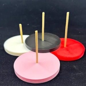 May include: A collection of colorful, round display stands. Each stand has a wooden dowel in the center. The stands are in various colors: white, black, red, and pink. The background is black.