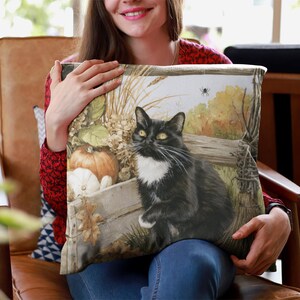 May include: A square throw pillow featuring a printed illustration of a black and white cat sitting next to a wooden crate filled with pumpkins and dried autumn foliage. A small spider hangs from a web in the upper corner of the scene.