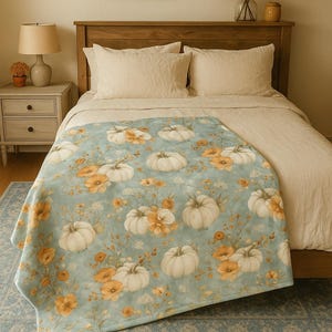 Puede incluir: Una manta azul claro con un estampado de calabazas blancas y flores naranjas. La manta está sobre una cama con almohadas blancas y una cabecera de madera. La habitación tiene un ambiente cálido y acogedor.