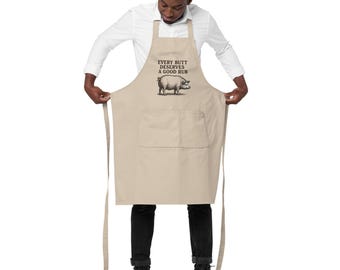 May include: A tan apron with a large front pocket and long ties. The apron features the text "EVERY BUTT DESERVES A GOOD RUB" above a black and white illustration of a pig. The apron is worn by a person.