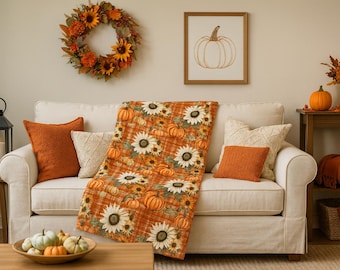 May include: A cozy living room scene with a white sofa adorned with orange pillows and a sunflower and pumpkin patterned throw blanket. A fall wreath and framed pumpkin art hang on the wall, with pumpkins and fall decor adding to the autumn theme.