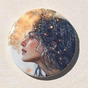 May include: A round, artistic button featuring a profile portrait of a person with dark hair and a celestial background. The portrait is detailed with soft, warm tones, and the background has a golden, sparkling effect.