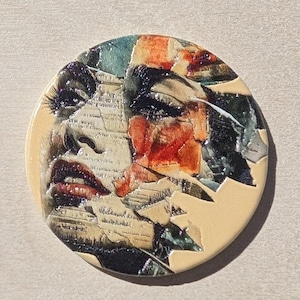 May include: Round ceramic art piece featuring a woman's face. The artwork uses a collage technique with fragments of text and colorful abstract elements. The woman's lips are a deep red, and the background is a light beige.