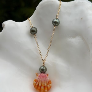 May include: A gold chain necklace featuring two dark grey pearls spaced along the links and a central pendant consisting of a single dark grey pearl attached to a small, fan-shaped seashell with vibrant orange and pink vertical stripes.