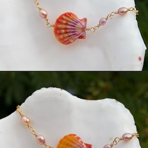 May include: A delicate bracelet featuring a central orange and pink scallop shell charm, accented with small, smooth, light pink oval beads. The components are connected by thin gold-toned wire links, creating a refined and symmetrical jewelry piece.