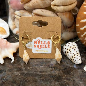 May include: Gold hoop earrings with small, cream-colored shell pendants. The earrings are displayed on a brown card with the text "Nells Shells" and "Provided by the Sea". The background features various seashells.