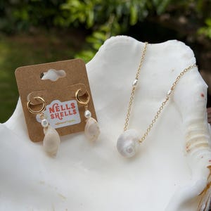 May include: A jewelry set featuring a gold chain necklace with a white seashell pendant and pearl accents, and matching gold hoop earrings with seashell and pearl drops. The earrings are displayed on a brown card with the text "NELLS SHELLS".