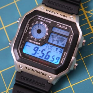 May include: A digital Casio watch with a black resin strap and a silver-tone case. The watch face shows the time, date, and a world map. The time reads 9:56:59 PM. Features include an illuminator and a 10-year battery.