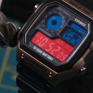 May include: A black and bronze Casio digital watch with a red digital display showing the time. The watch face includes blue and white accents, and the text "10 YEAR BATTERY" is visible. The watch has a black band.