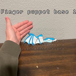 May include: A flat, paper-like finger puppet base featuring a blue and white dragon illustration with wings and a head. The text 'Finger puppet base 1' is printed at the top, and the watermark 'LilaCraftsPortland' is repeated across the image.