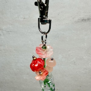 May include: A keychain featuring a miniature clear glass jar containing small green stems topped with translucent pink and red flower-shaped beads. The charm is attached to a silver-toned metal lobster clasp, creating a decorative accessory.