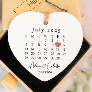 May include: A white heart-shaped ceramic ornament with a calendar for July 2025, highlighting the 11th. The names "Adam & Celeste" and the word "married" are below the calendar. The ornament is in a small box with a "Hand made" label.