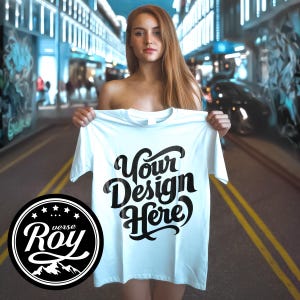 May include: White t-shirt with the text "Your Design Here" in a black, stylized font. The shirt is held up in front of a blurred urban background. A circular logo with the text "Roy" and mountain graphic is in the lower left corner.