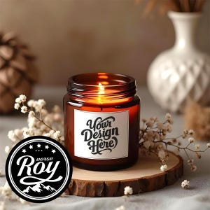 May include: A lit amber-colored candle in a glass jar with a white label that reads "Your Design Here." The candle sits on a wooden slice, surrounded by white flowers. A black and white logo that reads "Roy" is in the foreground.
