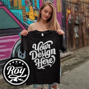 Puede incluir: Camiseta negra con el texto "Your Design Here" en escritura blanca. La camiseta se sostiene frente a un fondo de graffiti colorido. Un logotipo redondo con el texto "verse Roy" está en la esquina inferior izquierda.