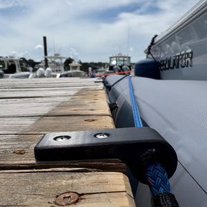 Op de afbeelding: Een zwarte bolder is vastgeschroefd aan een verweerde houten steiger. Een blauw en zwart touw is aan de bolder bevestigd en loopt naar een grijze boot. Andere boten zijn zichtbaar op de achtergrond onder een bewolkte lucht.