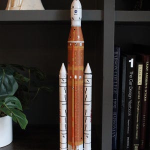 May include: A scale model of the NASA Space Launch System rocket featuring a tall, cylindrical orange core stage flanked by two white solid rocket boosters. The top section is white with a blue NASA logo and a pointed nose cone at the very peak.