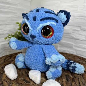 Può includere: Un gatto di peluche blu all'uncinetto con grandi occhi scintillanti. Il gatto ha segni blu scuro e una coda a fantasia. È seduto su una superficie tessuta con pietre bianche nelle vicinanze. Il giocattolo è fatto a mano.