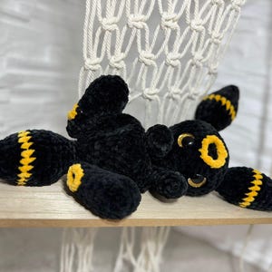 Könnte beinhalten: Ein schwarzes Häkeltier mit gelben Akzenten, das einem Zeichentrickcharakter ähnelt. Das Spielzeug hat große gelbe Augen, gelbe Streifen auf den Gliedmaßen und liegt auf einem Holzregal. Das Plüschtier ist weich und flauschig.
