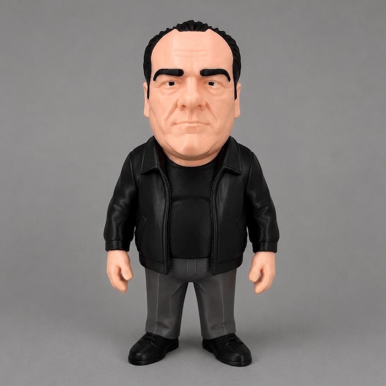 A detailed Tony Soprano collectible figure wearing a black leather jacket, black shirt, grey pants and black shoes. The figure is standing against a clean grey background.