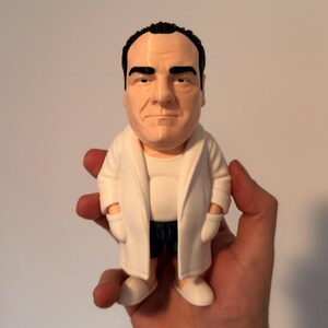 Peut inclure: Une figurine stylis&eacute;e d'une personne d'&acirc;ge m&ucirc;r aux cheveux fonc&eacute;s, portant un peignoir blanc sur un maillot de corps blanc et un short sombre. La figurine a le teint p&acirc;le, des sourcils &eacute;pais et une expression neutre, debout sur des chaussons blancs.