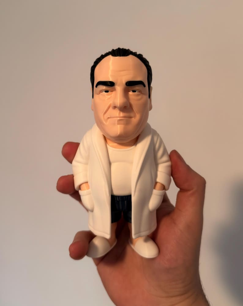 Peut inclure: Une figurine stylis&eacute;e d'une personne d'&acirc;ge m&ucirc;r aux cheveux fonc&eacute;s, portant un peignoir blanc sur un maillot de corps blanc et un short sombre. La figurine a le teint p&acirc;le, des sourcils &eacute;pais et une expression neutre, debout sur des chaussons blancs.