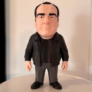 A detailed real life image of a Tony Soprano collectible figure wearing a black leather jacket, black shirt, grey pants and black shoes. The figure is standing on a white base against a white wall.