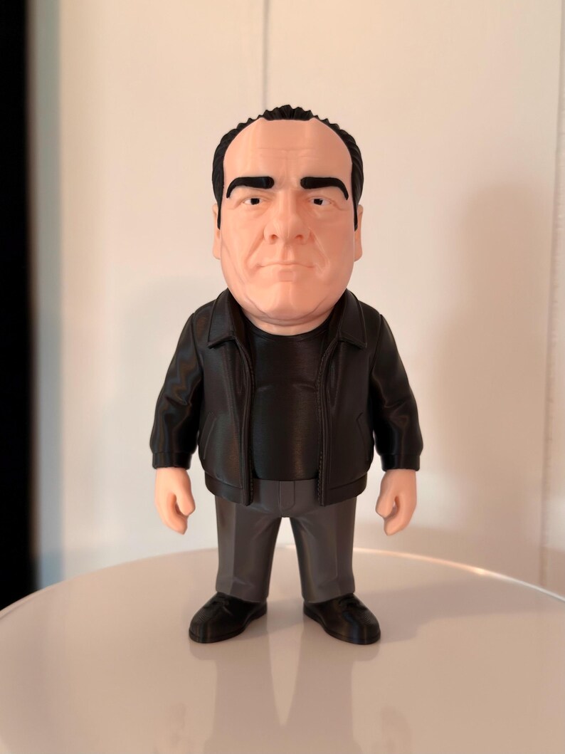 A detailed real life image of a Tony Soprano collectible figure wearing a black leather jacket, black shirt, grey pants and black shoes. The figure is standing on a white base against a white wall.