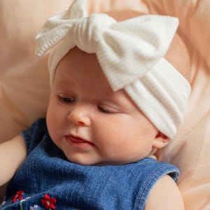 May include: A baby wearing a white, ribbed headband with a large bow. The baby is also wearing a sleeveless denim dress with embroidered flowers in red, white, and blue. The baby is lying on a peach-colored surface.