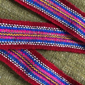 May include: A close-up of a red woven fabric ribbon with a colorful geometric pattern. The pattern includes blue, pink, yellow, and green threads.