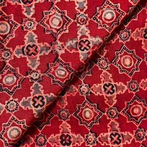 May include: A folded piece of fabric featuring a repeating geometric pattern in deep red, black, and muted beige. The design consists of interlocking star-shaped motifs and cross-like elements, creating a dense, symmetrical, and intricate visual texture.