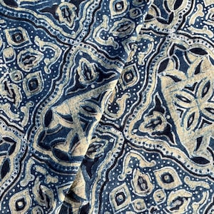 May include: Close-up of a fabric with a detailed, repeating pattern. The design features intricate shapes and floral motifs in shades of navy blue and beige. The fabric appears to be soft and flowing, with visible folds and creases.