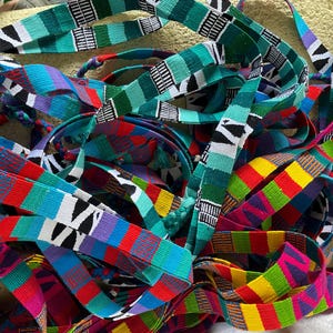 May include: A vibrant assortment of colorful woven fabric strips. The strips feature geometric patterns in shades of red, yellow, green, blue, and purple, with black and white accents. The textiles are likely used for crafting or decorative purposes.