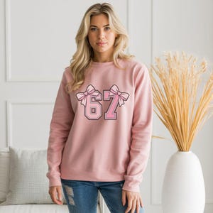 May include: A pale pink sweatshirt featuring the number "67" in a pink and black design, decorated with pink bows. The sweatshirt is a plain colour and has a crew neck.