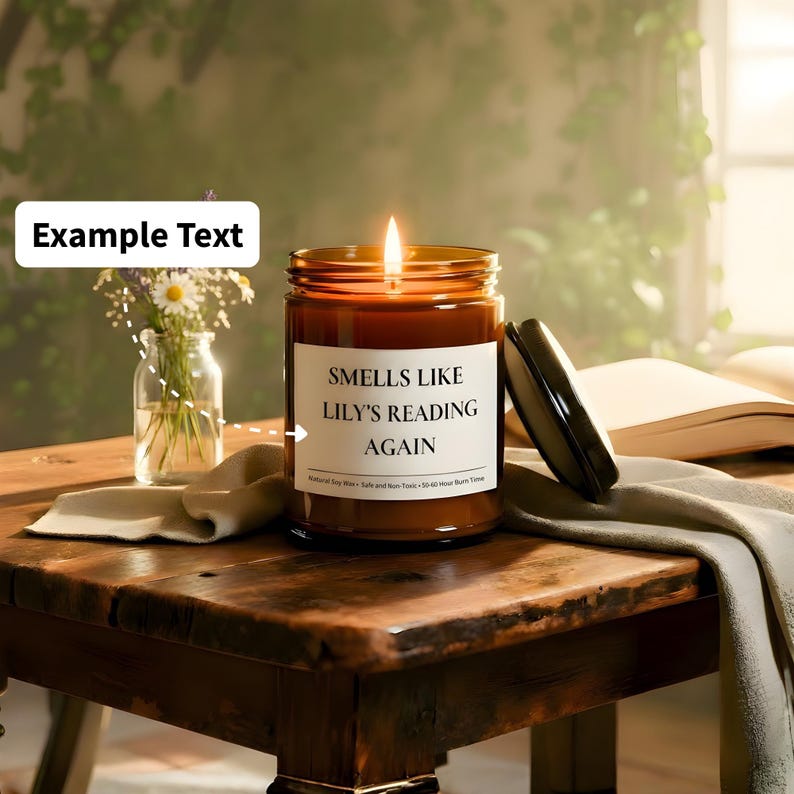 Puede incluir: Una vela encendida en un tarro de vidrio marr&oacute;n con el texto "Smells Like Lily's Reading Again" en una etiqueta blanca. La vela est&aacute; sobre una mesa de madera junto a un peque&ntilde;o jarr&oacute;n con flores y un libro abierto.