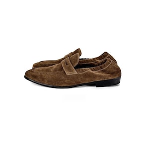 May include: A pair of brown suede loafers featuring a classic penny strap across the vamp, a rounded toe, and a low-profile black sole. The soft, textured leather upper has visible stitching along the edges and a flexible, unstructured silhouette.