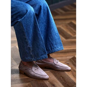 May include: Light grey suede loafers with a penny keeper detail. The shoes have a low heel and are worn with blue denim jeans. The loafers are a neutral colour, suitable for various outfits. The shoes are approximately 26 cm long.