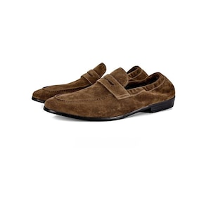 May include: A pair of brown suede loafers featuring a classic penny strap across the vamp, a rounded toe, and a low-profile black sole. The textured suede material covers the entire upper portion of the shoes, providing a uniform appearance.