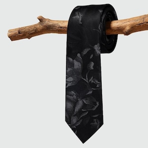Könnte beinhalten: Eine schwarze Krawatte mit einem grauen Blumenmuster hängt über einem Ast. Die Krawatte ist oben aufgerollt, das florale Design ist auf der Vorderseite sichtbar. Der Hintergrund ist einfarbig hellgrau.