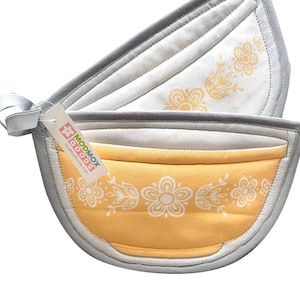May include: A pair of grey and yellow pot holders shaped like a teacup with a white floral design. The pot holders have a loop handle and a tag that reads "Modmox Goods".