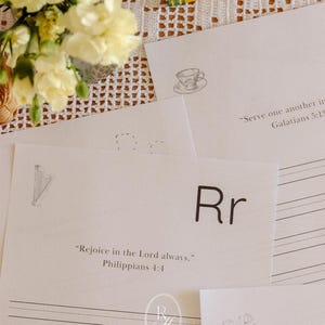 May include: A collection of white papers with handwritten text and illustrations, including a teacup, a harp, and a flower. The text includes the letter "Rr" and Bible verses. A bouquet of yellow flowers and a pencil are also visible.