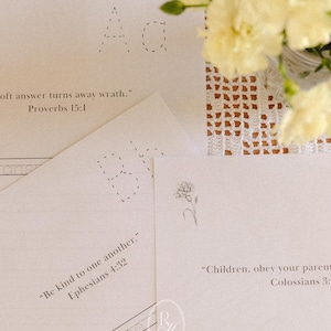 May include: A collection of cream-colored paper sheets with printed text and dotted letters. One sheet features the text "oft answer turns away wrath." Proverbs 15:1. Another sheet reads "Be kind to one another." Ephesians 4:32. A floral arrangement is in the background.
