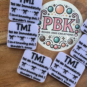 May include: A collection of square, glittery white resin pieces with the text "TMI Tell Me Immediately I love knowing shit I shouldn't know" in black. A round sticker with "PBK BEADS AND CREATIONS" is also visible.