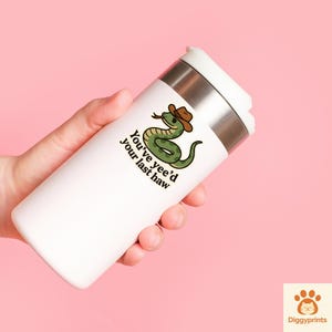 May include: White travel mug with a stainless steel band and white lid. The mug features a cartoon green snake wearing a brown cowboy hat with the text "You've yee'd your last haw."