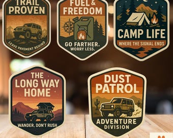 May include: A collection of five adventure-themed stickers with various designs. The stickers feature illustrations of a jeep, camping scenes, and desert landscapes, with text like "Trail Proven" and "Camp Life". The stickers are on a wooden surface.